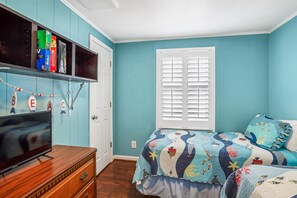 Quarto com parede verde-água, cama com colcha colorida, televisão sobre cômoda de madeira e janela com persianas.