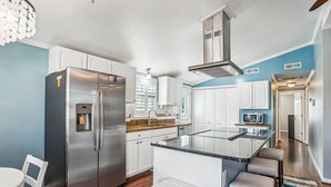 A modern kitchen with a stainless steel refrigerator, a built-in water dispenser, white cabinetry, a granite countertop, and a central island with seating. - Sea Glass Shack -- Pet Friendly with Hot Tub (Kill Devil Hills)