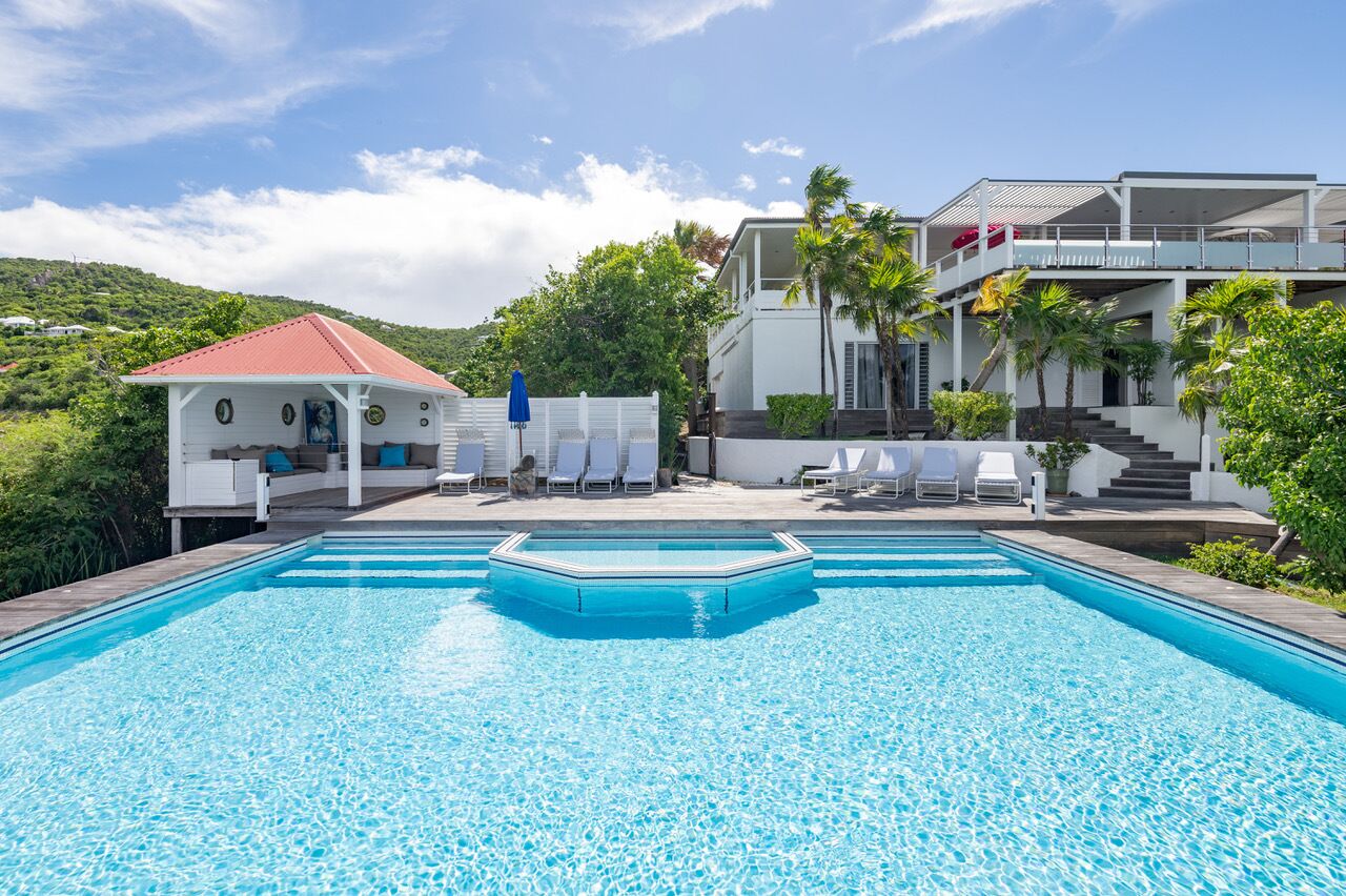 A swimming pool with lounge chairs and a covered area, surrounded by greenery and a hillside.