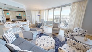 A modern living room with a grey sofa, patterned ottoman, and white armchairs. A dining area with a table and chairs is visible in the background.