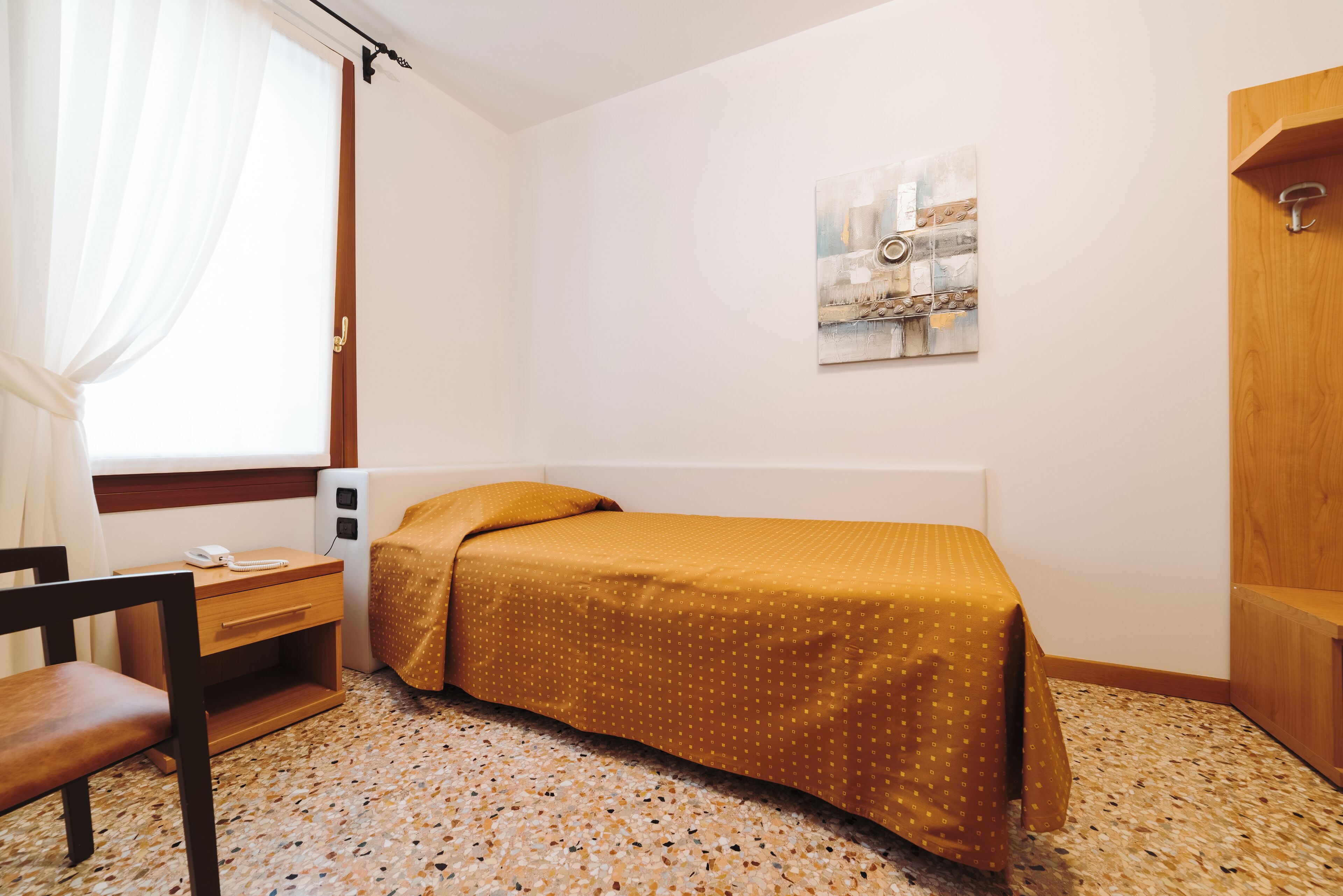 A small, neatly arranged bedroom with a bed, a chair, a bedside table with a phone, and a window with curtains.