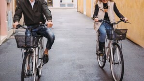 Deux personnes à vélo descendent une rue étroite, bordée de bâtiments aux fenêtres en arc de cercle et au toit recouvert de tuiles.