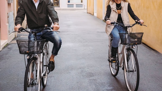 Dos personas andan en bicicleta por una calle estrecha bordeada de edificios con ventanas de arco y un tejado de baldosas.