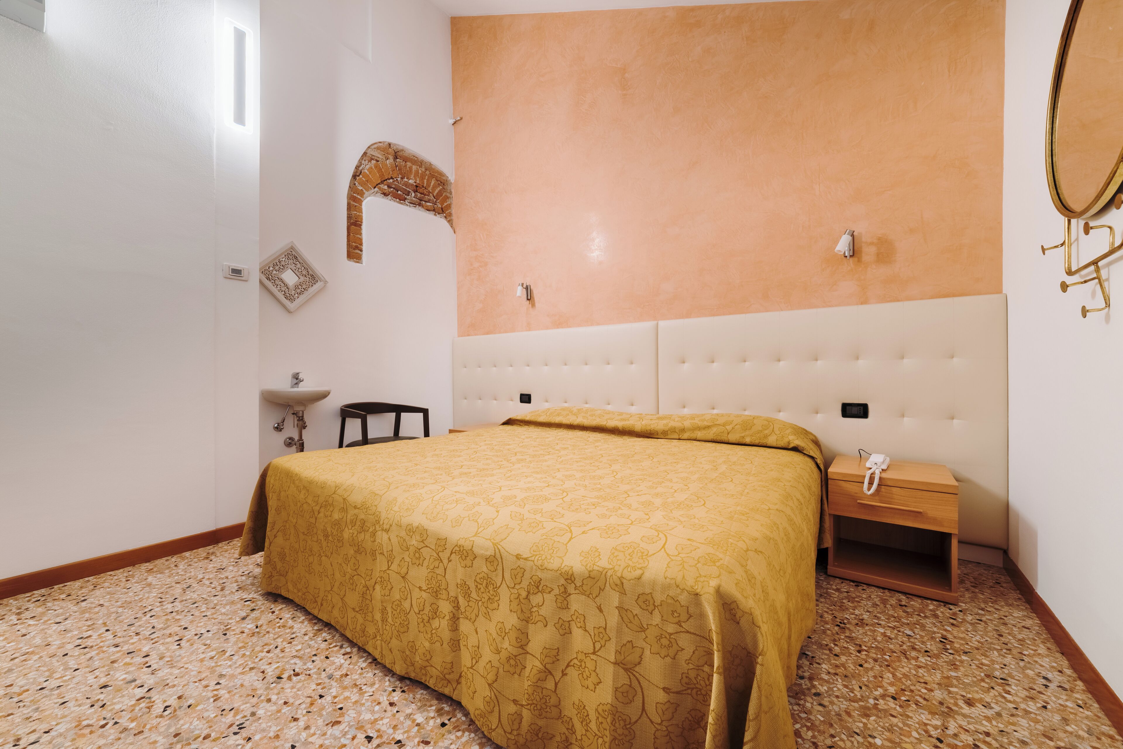 Ein Schlafzimmer mit einem Bett, einem Nachttisch, einem kleinen Tisch, einem Spiegel und einer wandmontierten Lampe.