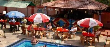 A swimming pool area with multiple umbrellas, chairs, and a tiled structure in the background.