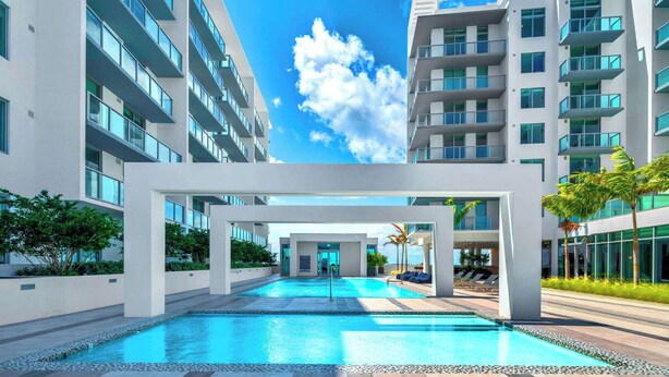 A modern hotel with a central swimming pool, multiple balconies, and a clear blue sky.