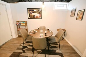 A dining area with a round table, four chairs, and a chandelier. There is a framed picture on the wall and a small shelf with books.