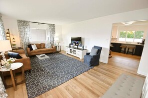 Sala de estar con sofá, mesa de centro y televisor, alfombra estampada y un área de cocina con taburetes.