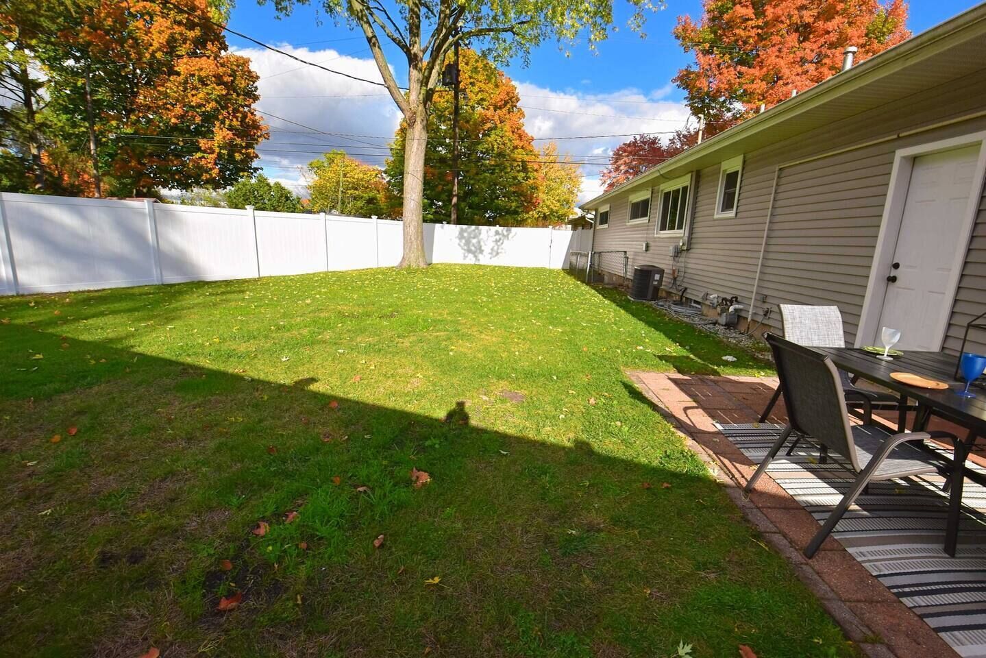 Un jardin avec une pelouse, une clôture blanche, un arbre et un espace patio comprenant une table et des chaises.