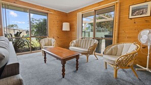 A room with a wooden interior, a large window, a sofa, two wicker chairs, a wooden coffee table, and a standing fan. - The Hideaway: This holiday house is your gateway to unforgettable memories (Paynesville)