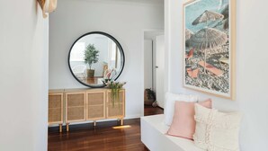 Un salón moderno con un sofá blanco, un espejo redondo, una consola de ratán y un cuadro con motivos playeros.