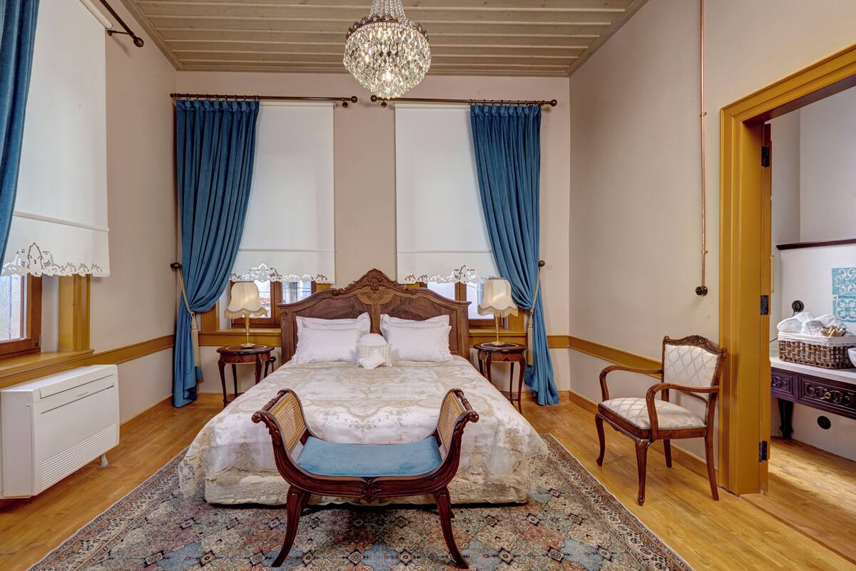 Un dormitorio amplio con una cama grande, dos mesitas de noche con lámparas, una silla de madera y una araña.
