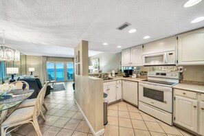 Eine moderne Küche mit weißen Schränken, einer Edelstahl-Mikrowelle und einem eingebauten Backofen. Es gibt eine Essecke mit einem Glastisch und Stühlen sowie einen Blick auf das Meer durch große Fenster.