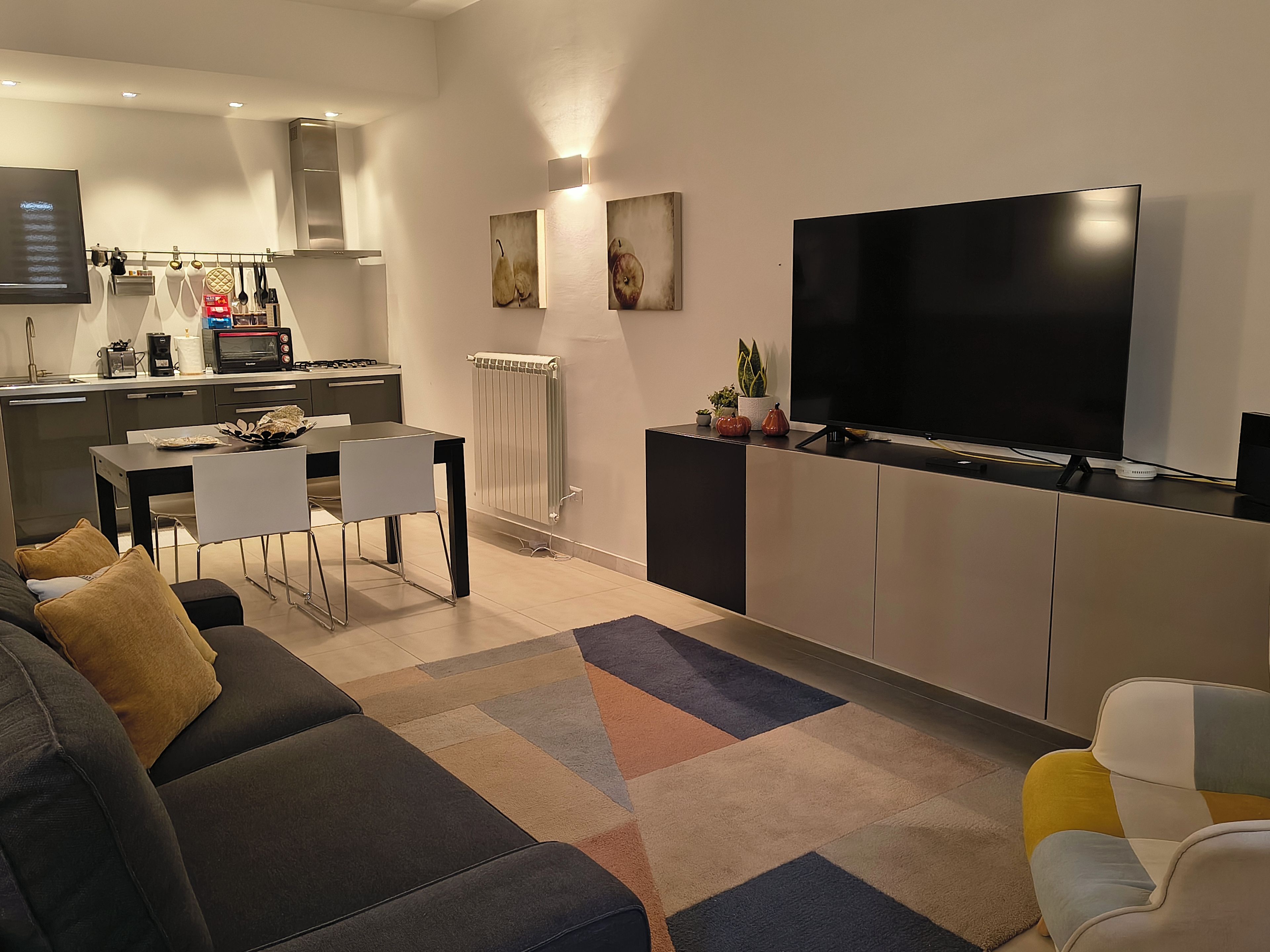 A modern living room with a sofa, a TV, a dining area with chairs, and a kitchen with appliances.