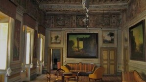 Et værelse med højt til loftet, detaljeret loftudsmykning, et stort maleri på væggen og et siddeområde med sofa og stole.