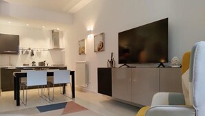 En moderne stue med et fladskærms-TV, et spiseområde med stole, og et køkken i baggrunden.