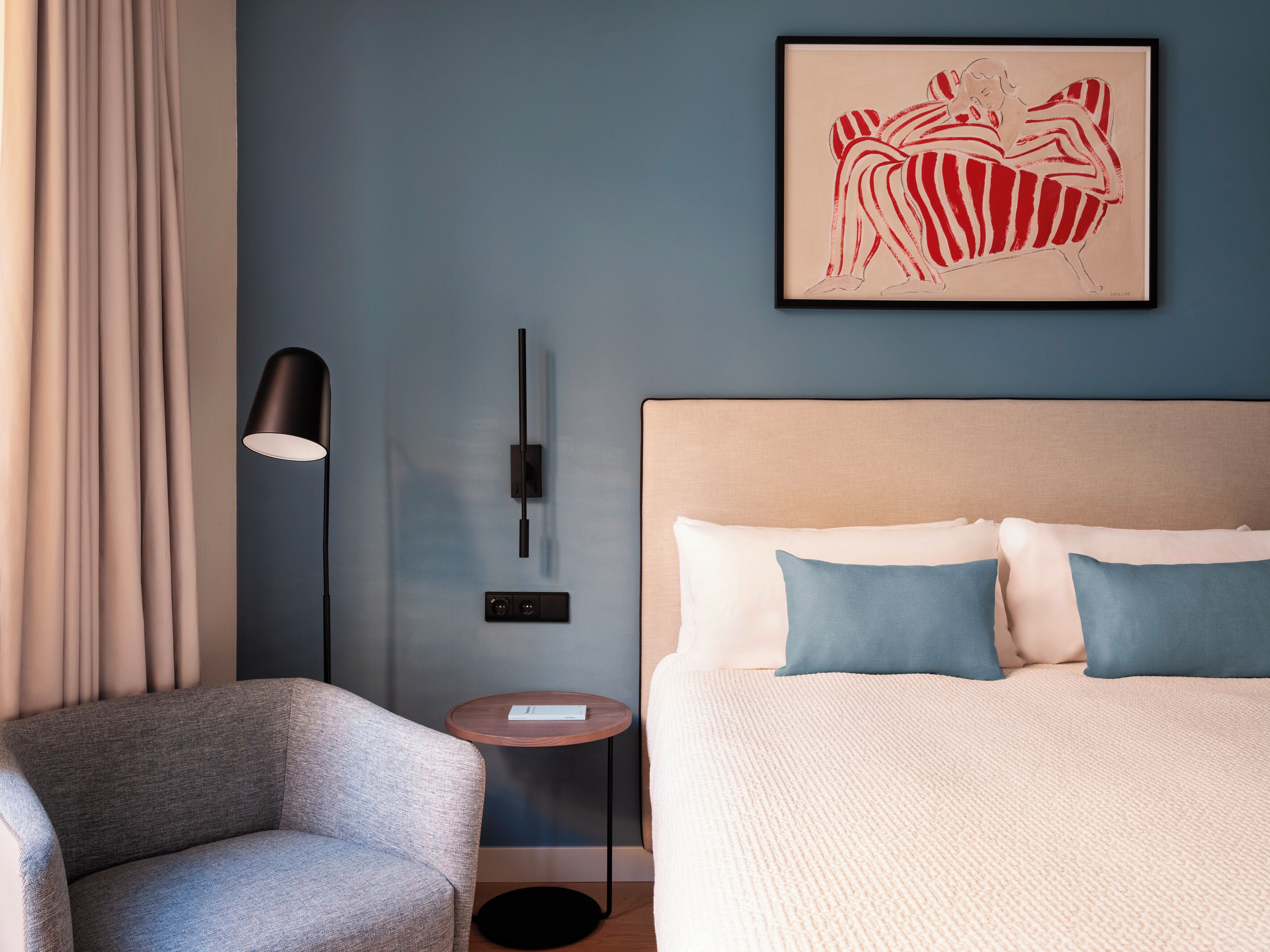 A modern hotel room with a bed, a chair, a side table, a lamp, and a wall art piece.