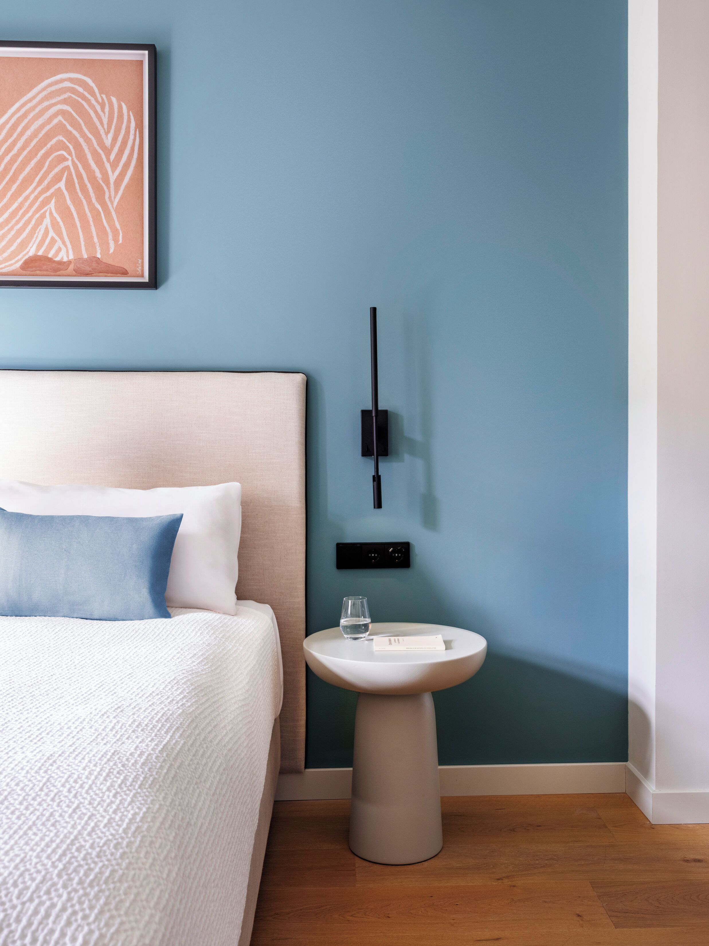 Une chambre à coucher avec un lit, une table de chevet sur laquelle se trouve un verre d’eau, et un luminaire fixé au mur.