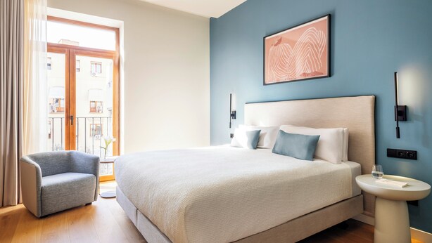 Una habitación de hotel moderna con una cama grande, una mesita de noche, una silla y vistas al exterior.
