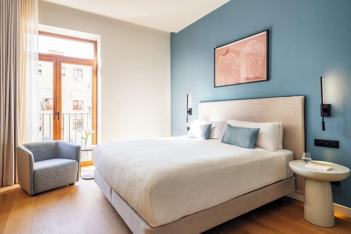 A modern hotel room with a large bed, a bedside table, a chair, and a view of the outside.