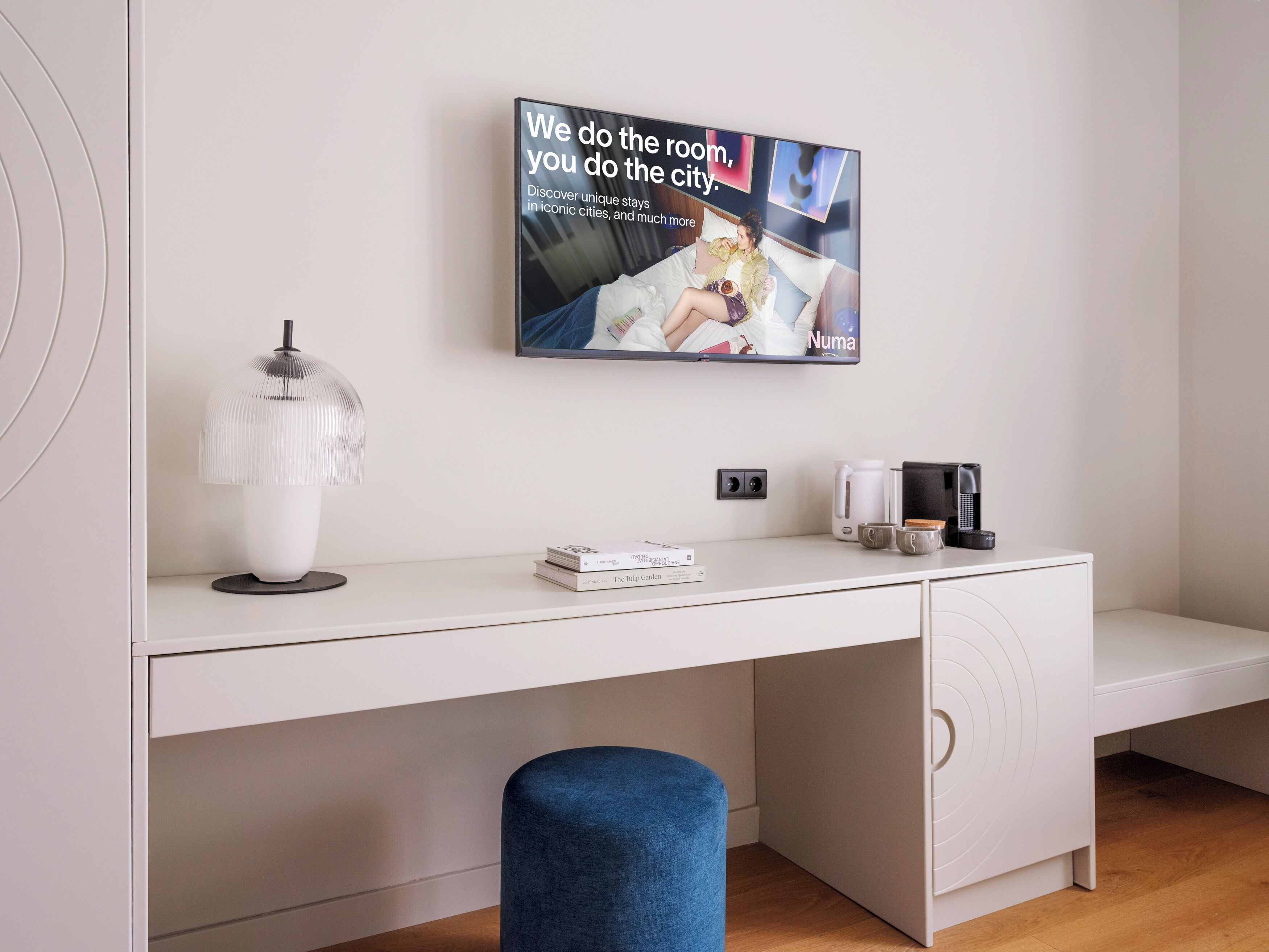 Una habitación moderna con un escritorio blanco, un televisor montado mostrando un anuncio, una cafetera y un reposapiés azul.