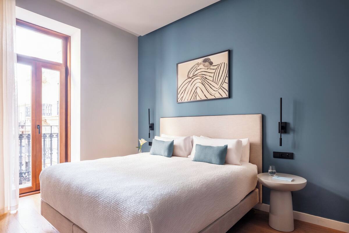 A bedroom with a large bed, a bedside table, a wall-mounted light, and a window with a view of buildings.