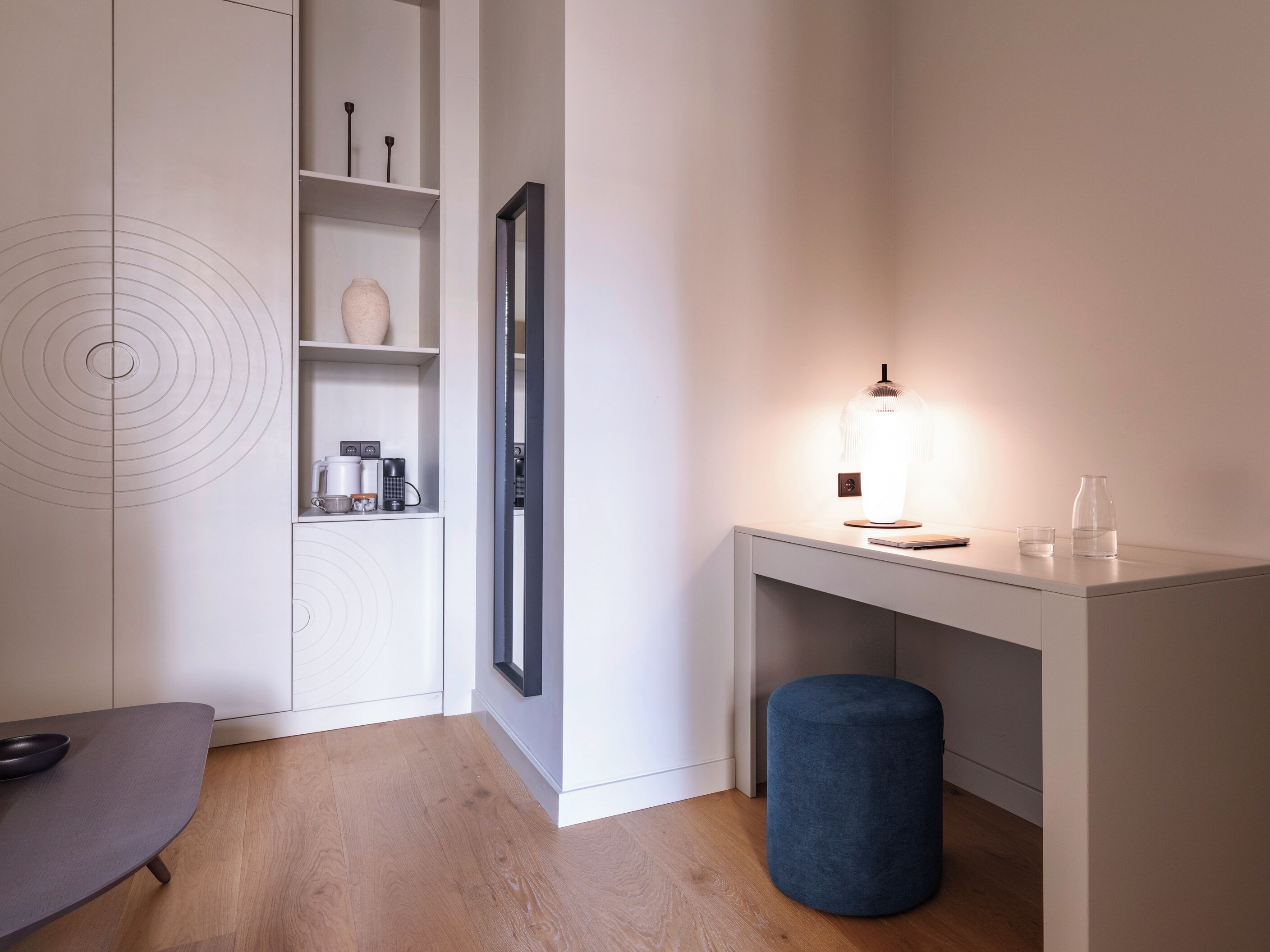 A modern, minimalist room with a white desk, a blue ottoman, and a shelf with coffee makers and a vase.