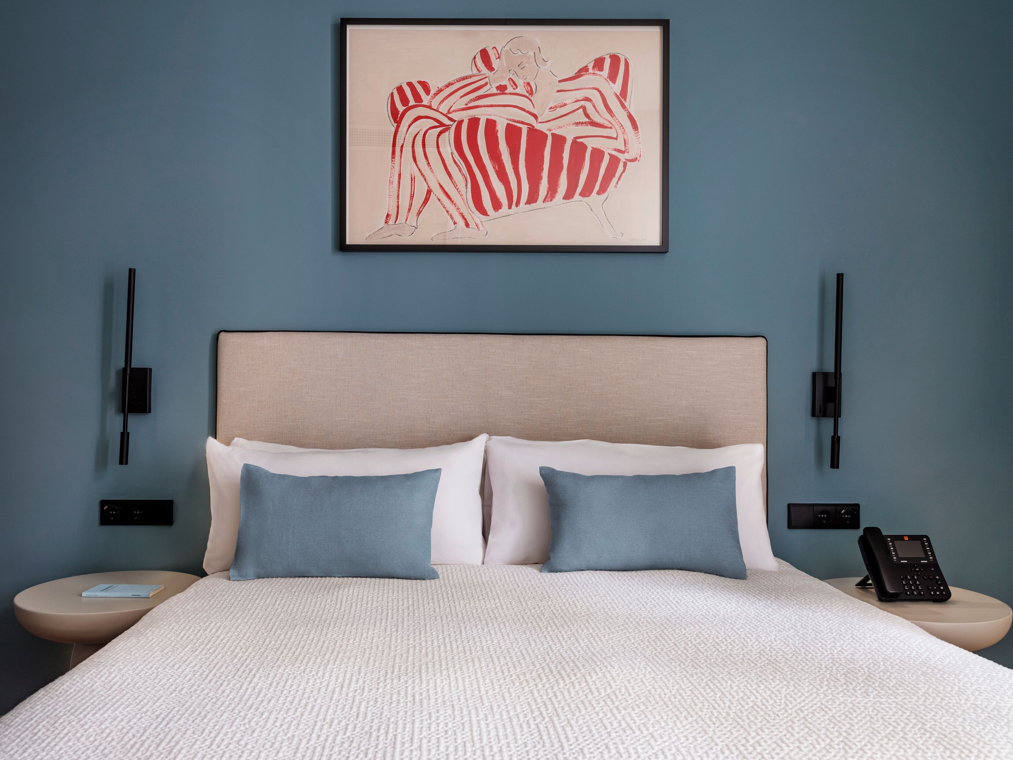 Habitació d'hotel amb un llit, dos coixins, una obra d'art emmarcada, taules de nit i un telèfon a la nit.