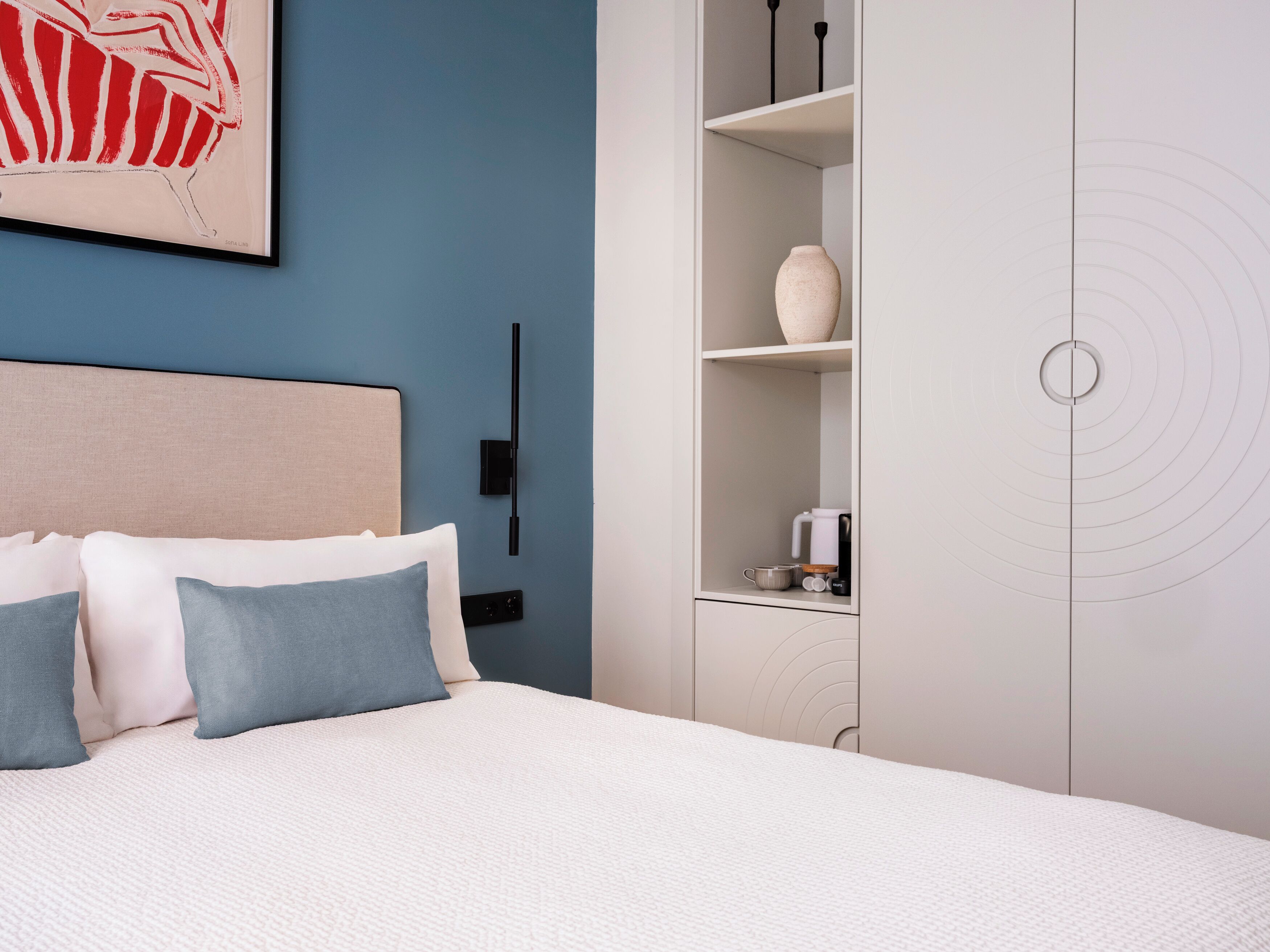 A bedroom with a bed, a nightstand, a wall-mounted lamp, a shelf with a vase, and a circular design on the wall.
