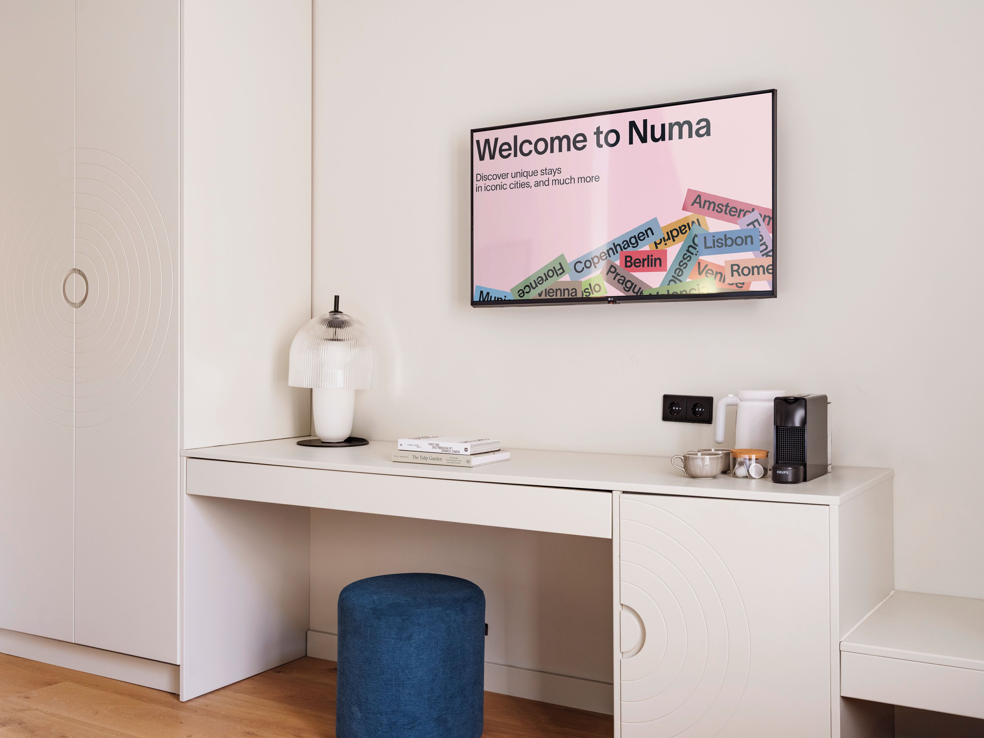 A modern, minimalist room with a white desk, a blue ottoman, a framed welcome sign, and a kettle.
