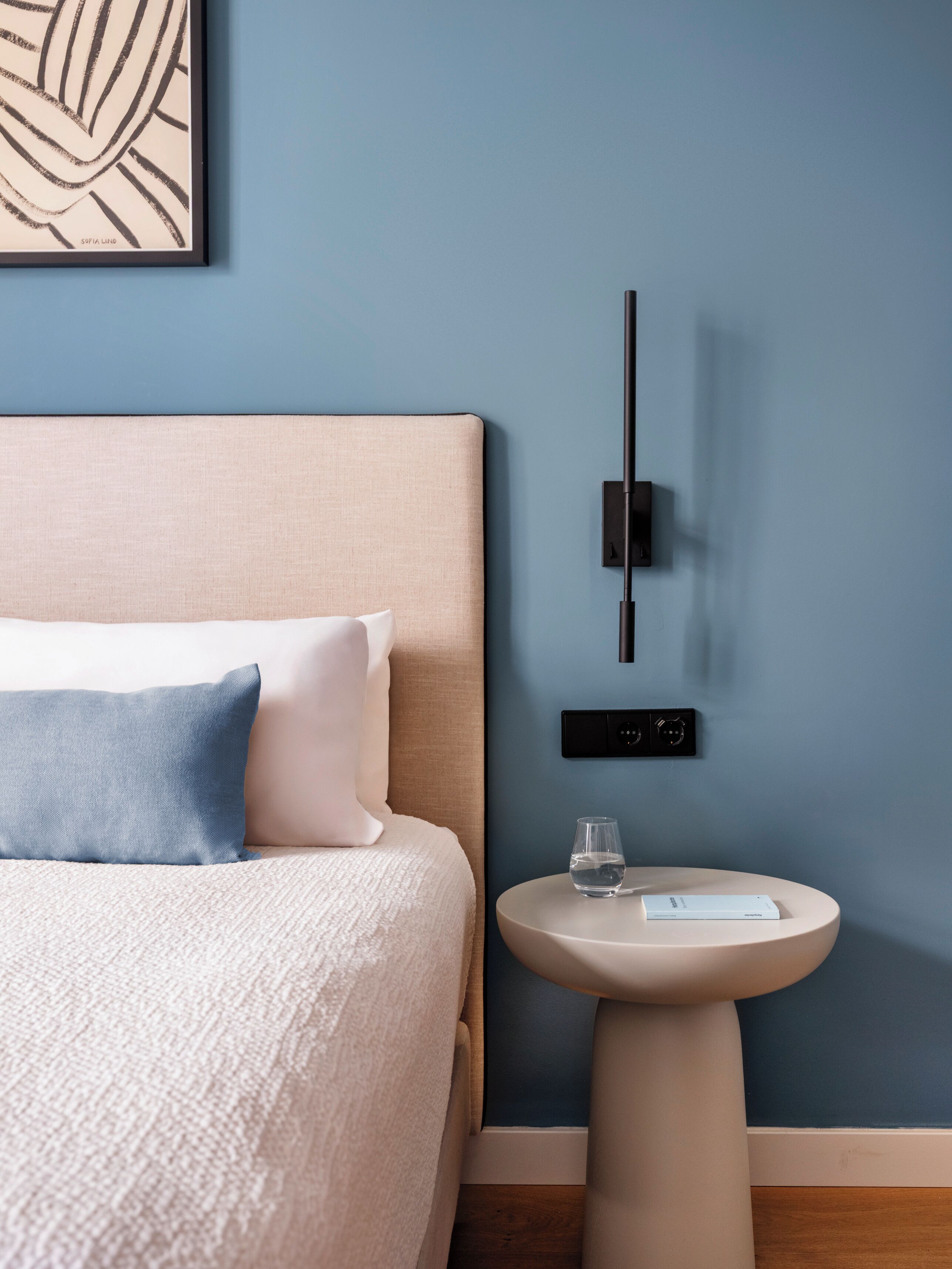 A neatly arranged bedroom with a bed, pillows, a bedside table with a glass of water, and a wall-mounted light fixture.