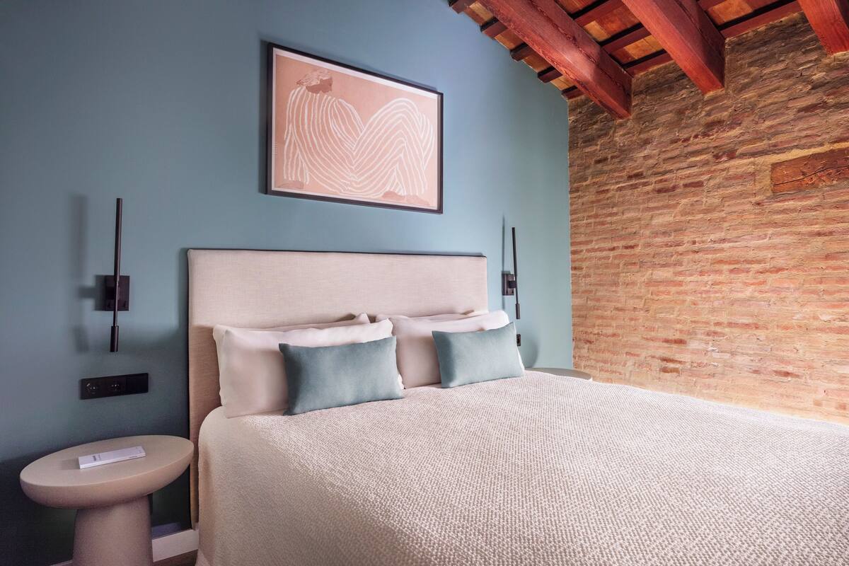 A bedroom with a bed, pillows, a bedside table, a wall-mounted light, and a framed artwork.