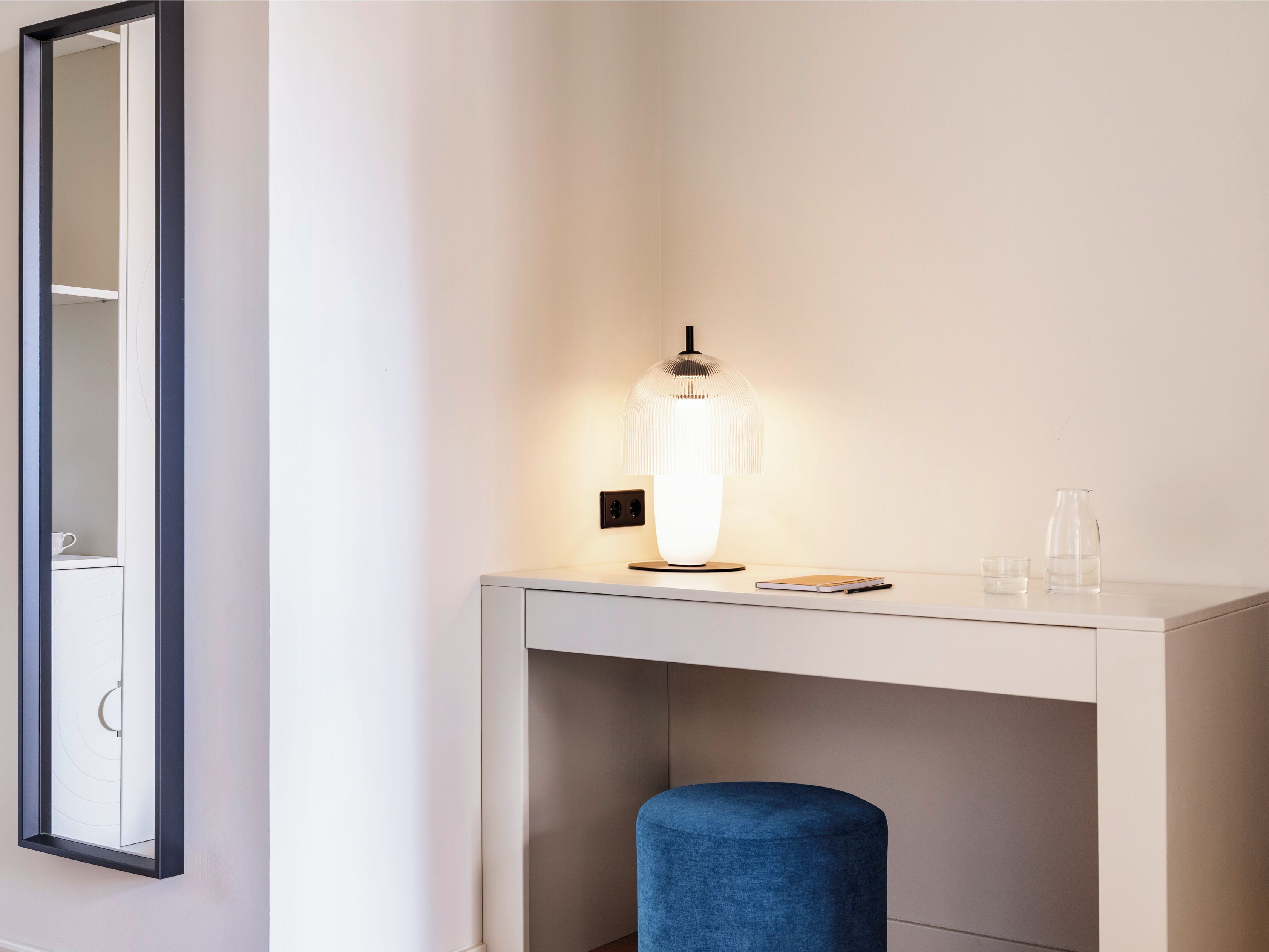 A white desk with a lamp, a blue ottoman, and a mirror on the wall.