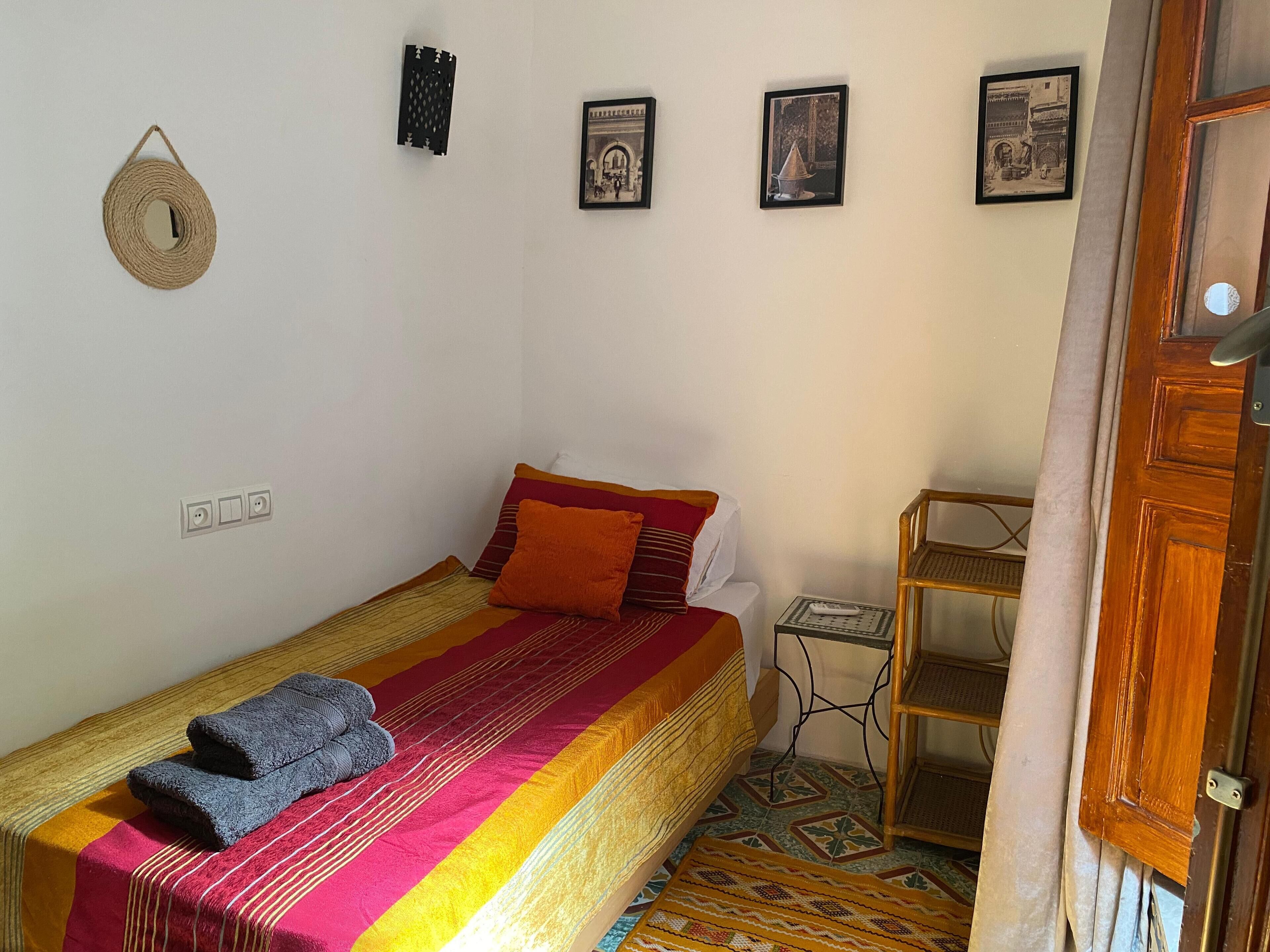A single bed with a colorful blanket and pillows, a small table with a remote control, and a wall-mounted hat.