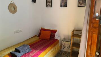 A single bed with a colorful blanket and pillows, a small table with a remote control, and a wall-mounted hat.