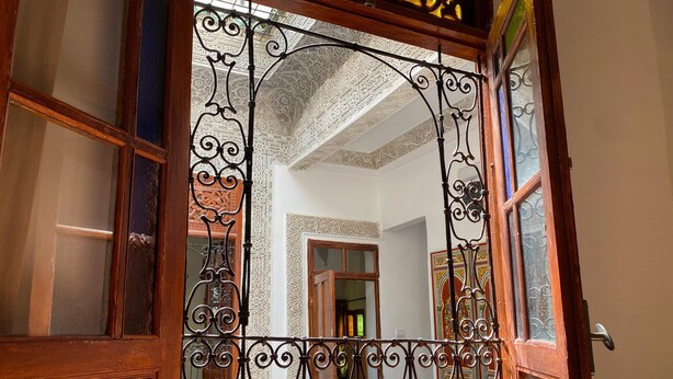 Un portail en fer forgé ouvragé, orné de motifs complexes, mène à une pièce dotée d’une porte en bois et offrant une vue sur un plafond à motifs et un escalier.