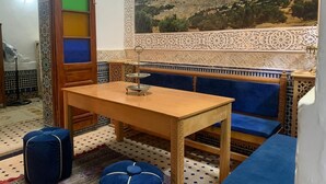 Une pièce avec une table en bois, des sièges rembourrés de couleur bleue, un mur orné d’un motif et un grand tableau représentant un paysage.
