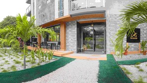 Un bâtiment moderne doté d’une entrée en verre, d’un espace extérieur aménagé et de plantes tropicales.