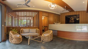 Un espace d’accueil avec un comptoir de réception en bois, deux fauteuils en osier, des ventilateurs de plafond et une grande fenêtre donnant sur un paysage verdoyant.