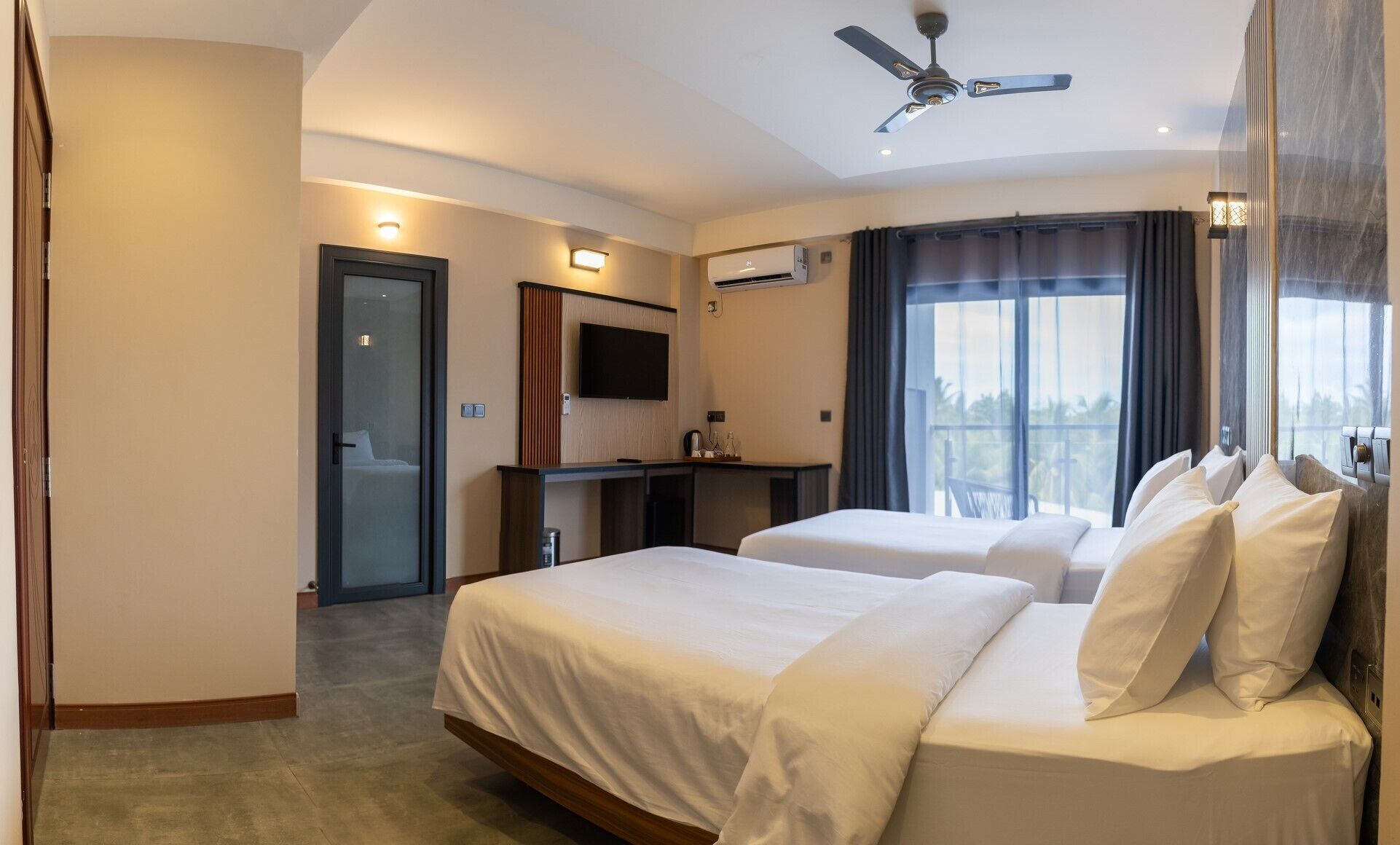 A hotel room with two beds, a desk, a flat-screen TV, an air conditioning unit, and a balcony with a view.