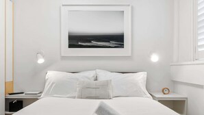 A neatly made bed with white linens, a framed picture of the ocean on the wall, a bedside lamp, and a small bedside table with a clock. - Bay View in Rushcutters Bay (Rushcutters Bay)