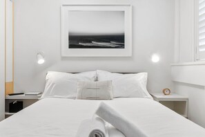 A neatly made bed with white linens, a framed picture of the ocean on the wall, a bedside lamp, and a small bedside table with a clock.