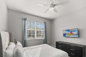 A bedroom with a bed, a ceiling fan, a window with curtains, a television on the wall, and a dark dresser.
