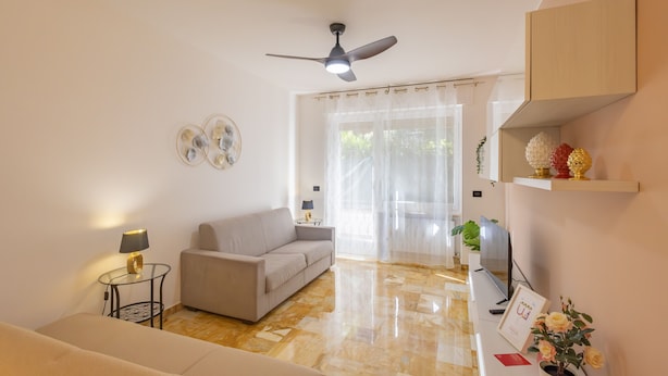 Un salon moderne comprenant un canapé, une petite table d’appoint avec une lampe, un ventilateur de plafond, un téléviseur et une porte donnant sur un balcon.