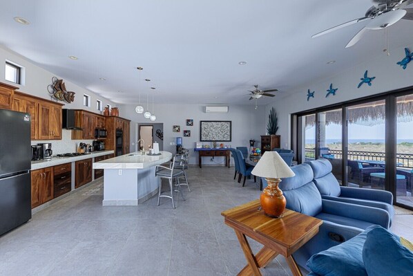 Una cocina moderna con una isla central, comedor con sillas azules y un área de estar con sofá y mesa.