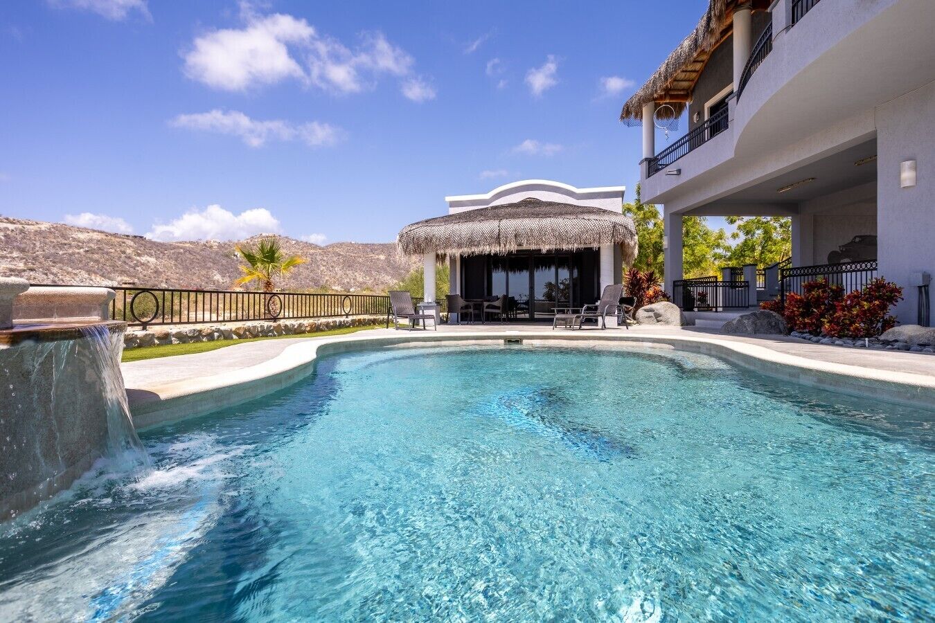 A swimming pool with a thatched-roof structure and lounge chairs, surrounded by a white building with a balcony.