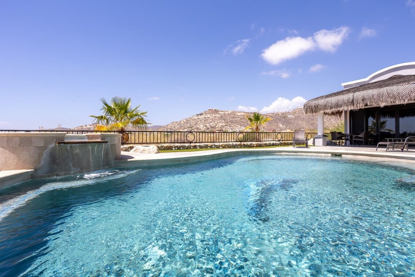 Una piscina cristallina con una struttura a tetto di paglia, palme e montagne sullo sfondo.