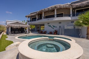 Un edificio de dos pisos con piscina, mobiliario exterior y una estructura con techo de paja.