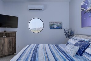 Un dormitorio con una cama, televisión, aire acondicionado, una ventana circular y un cuadro colgado en la pared.