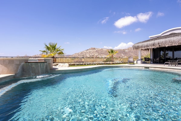 A swimming pool with clear water, a thatched-roof structure, palm trees, and a mountainous backdrop.